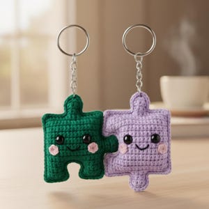 Può includere: Due portachiavi a forma di puzzle all'uncinetto. Uno è verde, l'altro è lavanda. Ognuno ha una faccia sorridente con occhi neri e guance rosa. I portachiavi sono collegati e appesi ad anelli argentati.