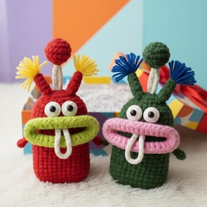 以下が含まれることがあります： 2つの編みぐるみのモンスターのおもちゃ。1つは赤色で、緑色の口、黄色のフリンジ、上に赤いボールが付いています。もう1つは緑色で、ピンク色の口、青色のフリンジ、上に緑色のボールが付いています。どちらも白い舌と大きな丸い目があります。