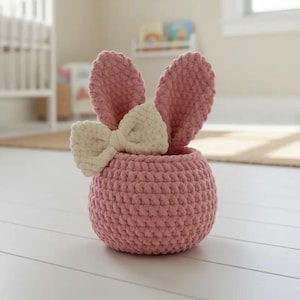 Peut inclure: Un panier rose en crochet fait à la main en forme de lapin. Le panier a deux oreilles droites et un nœud de couleur crème. Le panier est posé sur une surface blanche.