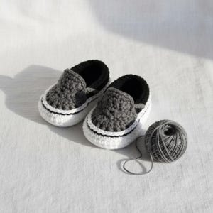 May include: A pair of handmade crochet baby shoes featuring a dark gray upper, a black collar, and a thick white sole with a black stripe detail. A small ball of matching gray yarn is positioned next to the shoes, showing the textured fiber construction.