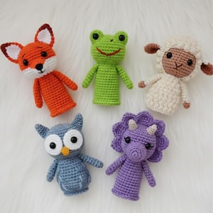 May include: Five handmade crochet finger puppets are displayed. The puppets include an orange and white fox, a green frog, a white sheep with brown details, a blue owl, and a purple triceratops dinosaur.