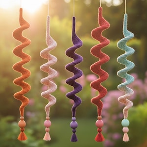 Può includere: Cinque decorazioni a spirale sospese, colorate in arancione, rosa chiaro, viola, rosso e turchese. Ognuna ha perline di legno e nappe sul fondo. Lo sfondo è una scena esterna sfocata con la luce del sole.