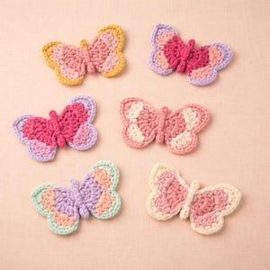 May include: A collection of six handmade crochet butterfly appliques in various pastel colors. Each butterfly features detailed wing patterns in shades of pink, purple, yellow, and green. The butterflies are arranged on a light pink surface.
