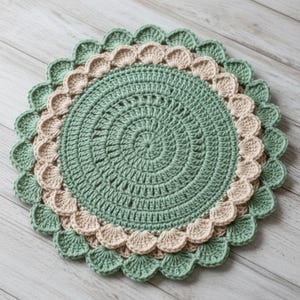 May include: A handmade, round crochet doily in shades of green and beige. The center is a solid green, with a spiral pattern. A beige scalloped border is surrounded by a green scalloped edge. The doily is placed on a light wood surface.