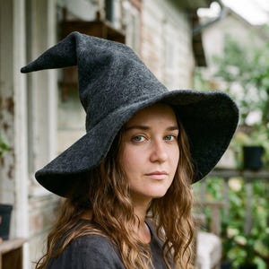 Puede incluir: Un sombrero de fieltro gris carbón oscuro con una copa alta y puntiaguda y un ala ancha ligeramente curvada. El sombrero presenta un acabado mate texturizado y una forma estructurada que se estrecha hasta una punta definida en la parte superior.
