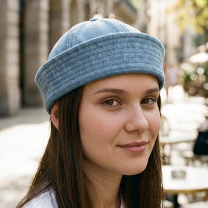 May include: A light blue denim docker cap featuring a rounded crown and a thick, multi-stitched rolled brim. The hat is constructed from panels of light-wash blue fabric with visible seams and a structured, brimless design that sits above the ears.
