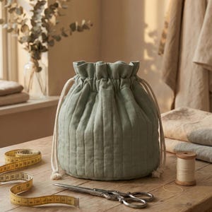 Puede incluir: Una bolsa verde salvia con cordón y líneas acolchadas verticales, sobre una mesa de madera. Una cinta métrica amarilla, tijeras vintage y una bobina de hilo están cerca. La bolsa tiene un cierre de cordón blanco.