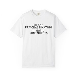 May include: White t-shirt with the text "im not procrastinating im doing SIDE QUESTS" in black. The shirt has a crew neck and short sleeves. The fabric appears to be a soft cotton blend.