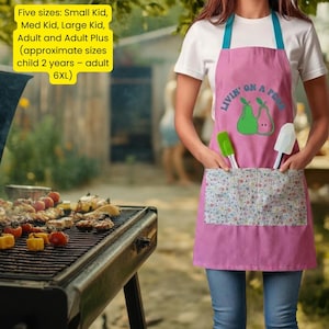 Puede incluir: Delantal rosa con correas azules y un bolsillo con estampado floral. El delantal tiene el texto "Livin' on a Pear" y un gráfico de dos peras. Una espátula verde y otra blanca están en el bolsillo.