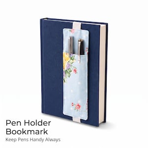 May include: A dark blue hardcover book featuring a light blue fabric pen holder bookmark attached with an elastic band. The bookmark displays a floral pattern with yellow, pink, and green flowers and white polka dots, holding two silver pens.