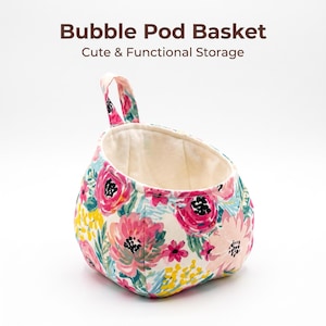 Op de afbeelding: Een stoffen opbergmand met een bloemenpatroon van roze, rode en gele bloemen op een witte achtergrond. De mand heeft één handvat aan de bovenkant en de tekst Bubble Pod Basket Cute & Functional Storage staat erboven gedrukt.