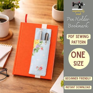 May include: A light blue pen holder bookmark with a floral pattern, holding two pens, is inserted into an orange book. The bookmark has a white polka dot design and a red ribbon. The text on the image says "Pen Holder Bookmark" and "One Size".