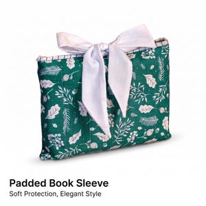 May include: A rectangular padded book sleeve featuring a dark green fabric with a white botanical print of leaves, pine needles, and berries. The sleeve is secured at the top with a large, tied white fabric bow and has vertical quilted stitching.