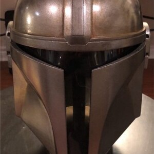 Puede incluir: Un casco Beskar de Mandaloriano plateado con un acabado desgastado. El casco tiene una visera en forma de T y una cresta en el centro. El texto "THE MANDALORIAN - BESKAR HELMET" est&aacute; en una placa plateada debajo del casco. La placa tambi&eacute;n incluye el texto "WEIGHT 2.72 KG" y "WIDTH 240 MM".