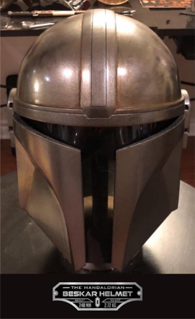 Puede incluir: Un casco Beskar de Mandaloriano plateado con un acabado desgastado. El casco tiene una visera en forma de T y una cresta en el centro. El texto "THE MANDALORIAN - BESKAR HELMET" est&aacute; en una placa plateada debajo del casco. La placa tambi&eacute;n incluye el texto "WEIGHT 2.72 KG" y "WIDTH 240 MM".