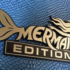 May include: A metallic emblem featuring a stylized mermaid silhouette with a flowing tail and hair, positioned above a rectangular black plaque with the white text "MERMAID EDITION" embossed on a textured, bright blue background surface.