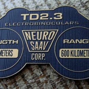 May include: A blue and gold electrobinoculars device with the text "TD2.3 ELECTROBINOCULARS" and "NEURO SAAV CORP." The device has a range of 600 kilometres and a length of 0.25 metres.