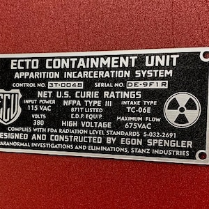 Peut inclure: Une plaque en métal noir et argent avec le texte "ECTO CONTAINMENT UNIT APPARITION INCARCERATION SYSTEM" et "NET U.S. CURIE RATINGS". La plaque comprend également un symbole de radiation et le texte "DESIGNED AND CONSTRUCTED BY EGON SPENGLER".