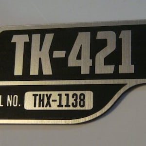 May include: A silver metal plate with black lettering. The plate has a serial number, "THX-1138", and a model number, "TK-421".