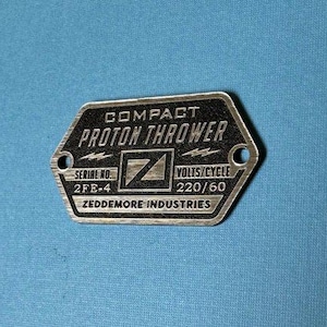 Peut inclure: Plaque métallique argentée et noire avec les mots "COMPACT PROTON THROWER" et "ZEDDEMORE INDUSTRIES". La plaque comporte deux trous et le texte "SERIAL NO. 2FE-4" et "VOLTS/CYCLE 220/60".