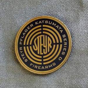 May include: A gold and black round sticker with the text "Steyr Pflager Katsumata Series D Firearms" around a black and gold target with the word "Steyr" in the centre.