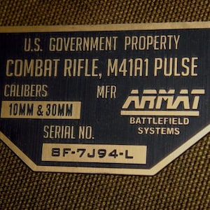 Pode incluir: Uma placa de metal preto com guarnição dourada, etiquetada como "U.S. GOVERNMENT PROPERTY COMBAT RIFLE, M41A1 PULSE". A placa também inclui os calibres "10MM & 30MM", o fabricante "ARMAT BATTLEFIELD SYSTEMS" e o número de série "BF-7J94-L".