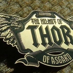 May include: A gold and black metal emblem with a winged design. The text reads "The Helmet of Thor of Asgard".