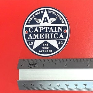 Pu&ograve; includere: Un adesivo rotondo blu e bianco con una stella e il testo "WORLD WAR II LIVING LEGEND CAPTAIN AMERICA 19 THE FIRST AVENGER 42".