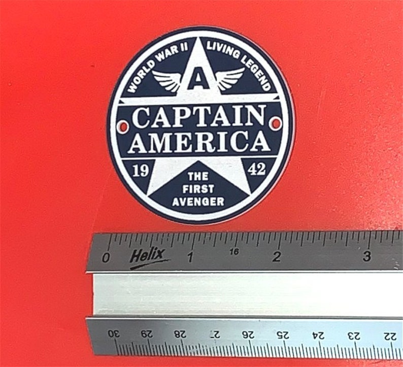 Pu&ograve; includere: Un adesivo rotondo blu e bianco con una stella e il testo "WORLD WAR II LIVING LEGEND CAPTAIN AMERICA 19 THE FIRST AVENGER 42".