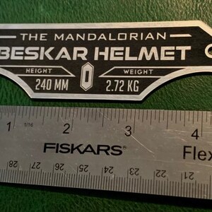 Puede incluir: Una placa de metal plateado con texto negro que dice "The Mandalorian - Beskar Helmet". La placa tambi&eacute;n incluye el texto "Altura 240 mm" y "Peso 2,72 kg".