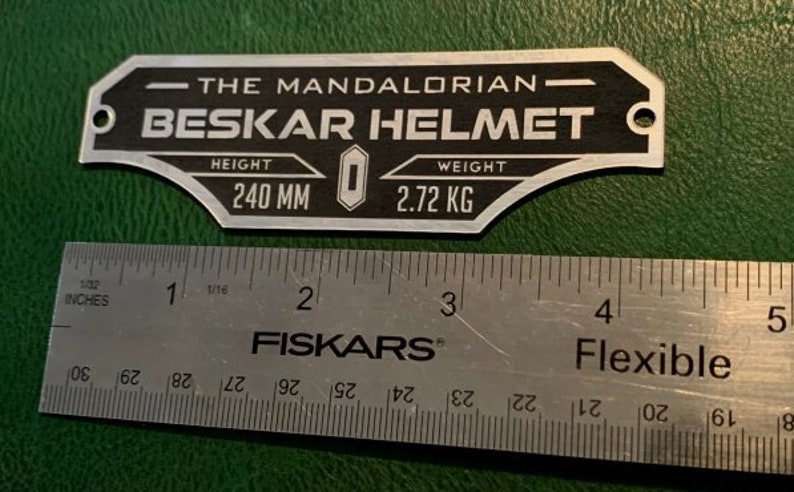 Puede incluir: Una placa de metal plateado con texto negro que dice "The Mandalorian - Beskar Helmet". La placa tambi&eacute;n incluye el texto "Altura 240 mm" y "Peso 2,72 kg".