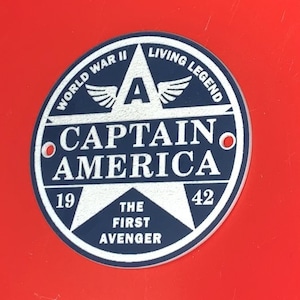 Pu&ograve; includere: Un adesivo circolare blu e bianco con una stella al centro. La stella ha il testo "Captain America" in lettere bianche. L'adesivo ha anche il testo "World War II" e "Living Legend" attorno alla stella. Il testo "The First Avenger" &egrave; nella parte inferiore della stella.