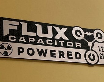 May include: Black and silver sticker with the text "Flux Capacitor Powered" and a stylized illustration of a circuit board with a radiation symbol and the text "1.21 GW".