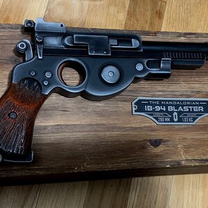 May include: A black and silver replica of the Mandalorian's  'The Mandalorian'  IB-94 Blaster, mounted on a wooden display stand. The display stand has a metal plaque with the text 'THE MANDALORIAN IB-94 BLASTER LENGTH 260 MM WEIGHT 1.03 KG'.