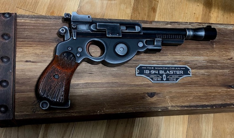 May include: A black and silver replica of the Mandalorian's  'The Mandalorian'  IB-94 Blaster, mounted on a wooden display stand. The display stand has a metal plaque with the text 'THE MANDALORIAN IB-94 BLASTER LENGTH 260 MM WEIGHT 1.03 KG'.