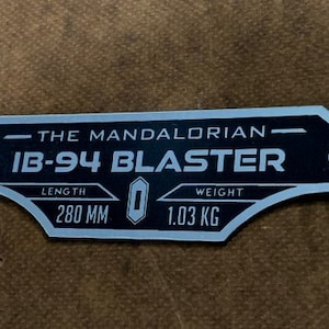 May include: A silver metal plaque with black text that reads "The Mandalorian - IB-94 Blaster". The plaque also includes the text "Length 280 MM" and "Weight 1.03 KG".