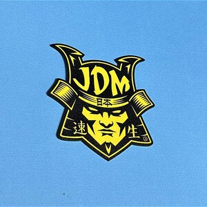 May include: Black and yellow sticker with a samurai warrior wearing a helmet with the word "JDM" in yellow letters and the word "日本" in Japanese characters above the warrior's face. The words "速生" are on the left and right sides of the warrior's face.