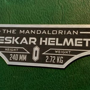 Puede incluir: Una placa de metal plateado con texto negro que dice "The Mandalorian Beskar Helmet". La placa tiene dos agujeros en la parte superior e inferior. La placa tambi&eacute;n tiene el texto "Height 240 MM" y "Weight 2.72 KG".