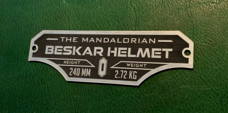 Puede incluir: Una placa de metal plateado con texto negro que dice "The Mandalorian Beskar Helmet". La placa tiene dos agujeros en la parte superior e inferior. La placa tambi&eacute;n tiene el texto "Height 240 MM" y "Weight 2.72 KG".