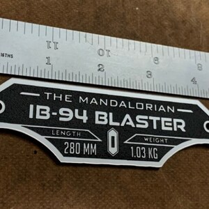 May include: A black and silver metal plaque with the text "The Mandalorian IB-94 Blaster" and the specifications "Length 280 MM Weight 1.03 KG".