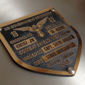 Puede incluir: Una placa de metal dorada con texto negro. La placa tiene forma de escudo y tiene un pequeño águila con las letras "SSR" en su pecho. El texto en la placa dice "US Government Property", "Control No. 030262-JW", "Serial No. SK-031941", "Combat Shield, Vibrantum", "Issued to Capt. Steve Rogers", "Mfr Stark Industries", "Strategic Scientific Reserve".