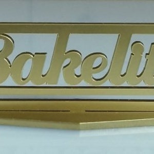 May include: A gold-colored metal sign with the word "Bakelite" cut out in the center. The sign has a decorative design on the ends.