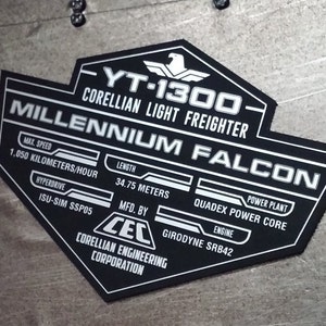 Puede incluir: Una placa de metal negro y plateado con el texto "YT-1300 Corellian Light Freighter Millennium Falcon" y especificaciones técnicas, incluyendo "Velocidad máxima 1.050 kilómetros/hora", "Longitud 34,75 metros", "Hyperdrive ISU-SIM SSP05", "Planta de energía Quadez Power Core", "Motor Girodyne SRB42", y "Fabricada por CEC Corellian Engineering Corporation".