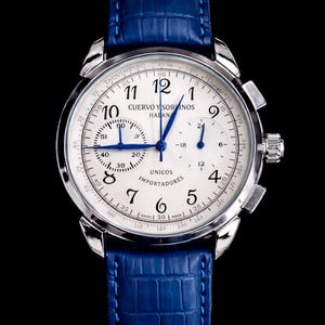 May include: A silver wristwatch with a white face and blue hands, featuring the text "CUERVO Y SOBRINOS". The watch has a blue leather band and a detailed dial with multiple sub-dials and markings.