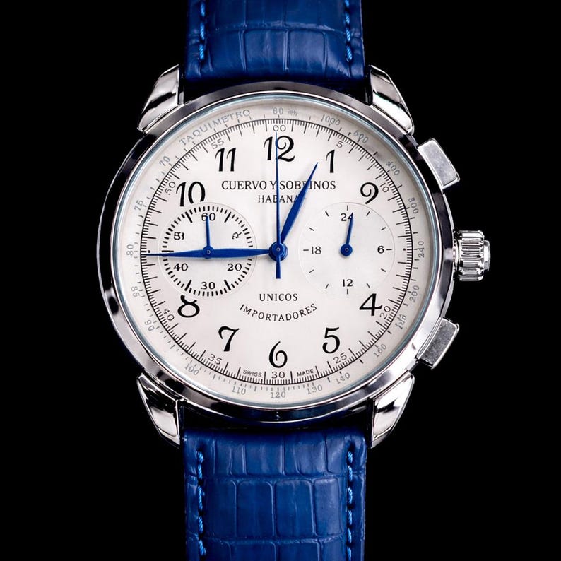 May include: A silver wristwatch with a white face and blue hands, featuring the text "CUERVO Y SOBRINOS". The watch has a blue leather band and a detailed dial with multiple sub-dials and markings.