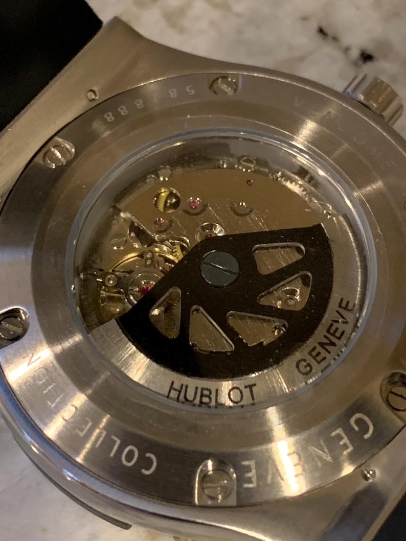 May include: Close-up of the back of a Hublot watch, showcasing the intricate internal mechanisms. The watch features a transparent window revealing the gears, jewels, and rotor. The words "Hublot" and "Geneve" are visible on the watch.