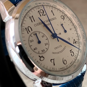 May include: A silver-toned wristwatch with a cream-colored face and blue hands. The watch features multiple sub-dials and the text "CUERVOS Y SORINOS HABANOS". The watch has a blue leather band.