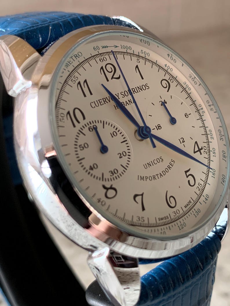 May include: A silver-toned wristwatch with a cream-colored face and blue hands. The watch features multiple sub-dials and the text "CUERVOS Y SORINOS HABANOS". The watch has a blue leather band.