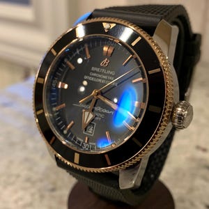 Może przedstawiać: Zegarek automatyczny Breitling Superocean z czarną tarczą, złotymi akcentami i czarnym gumowym paskiem. Zegarek posiada obrotową lunetę i wyświetlacz daty. Zegarek jest prezentowany na drewnianym stojaku.