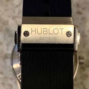 May include: Close-up of a luxury watch clasp. The silver clasp features the engraved brand name "HUBLOT GENEVE" and secures a black ribbed rubber strap. The watch band is dark blue on the inside.